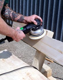 Tätowierte Hände eines Mannes halten eine Schleifmaschine und bearbeiten eine Holzbank im Rahmen des Holzbearbeitungslehrgang